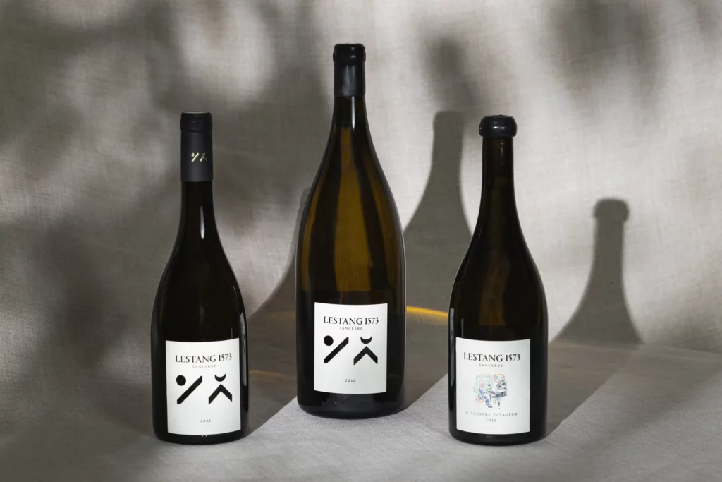 Trois bouteilles de vin Lestang 1573 millésime 2022 (format standard et magnum), posées sur un tissu beige, éclairées par une lumière douce qui projette leur ombre sur le fond.