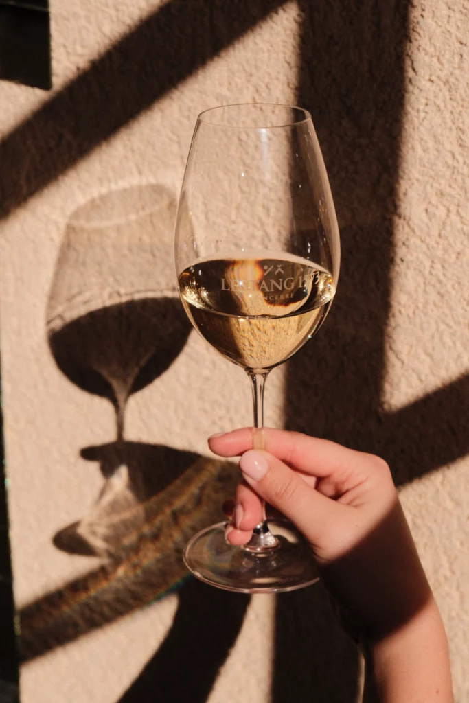 Une main féminine tient un verre à pied rempli de vin blanc Lestang 1573 devant un mur texturé. On distingue le logo “Lestang 1573” gravé sur le verre et le jeu d’ombres portées sur le mur derrière.
